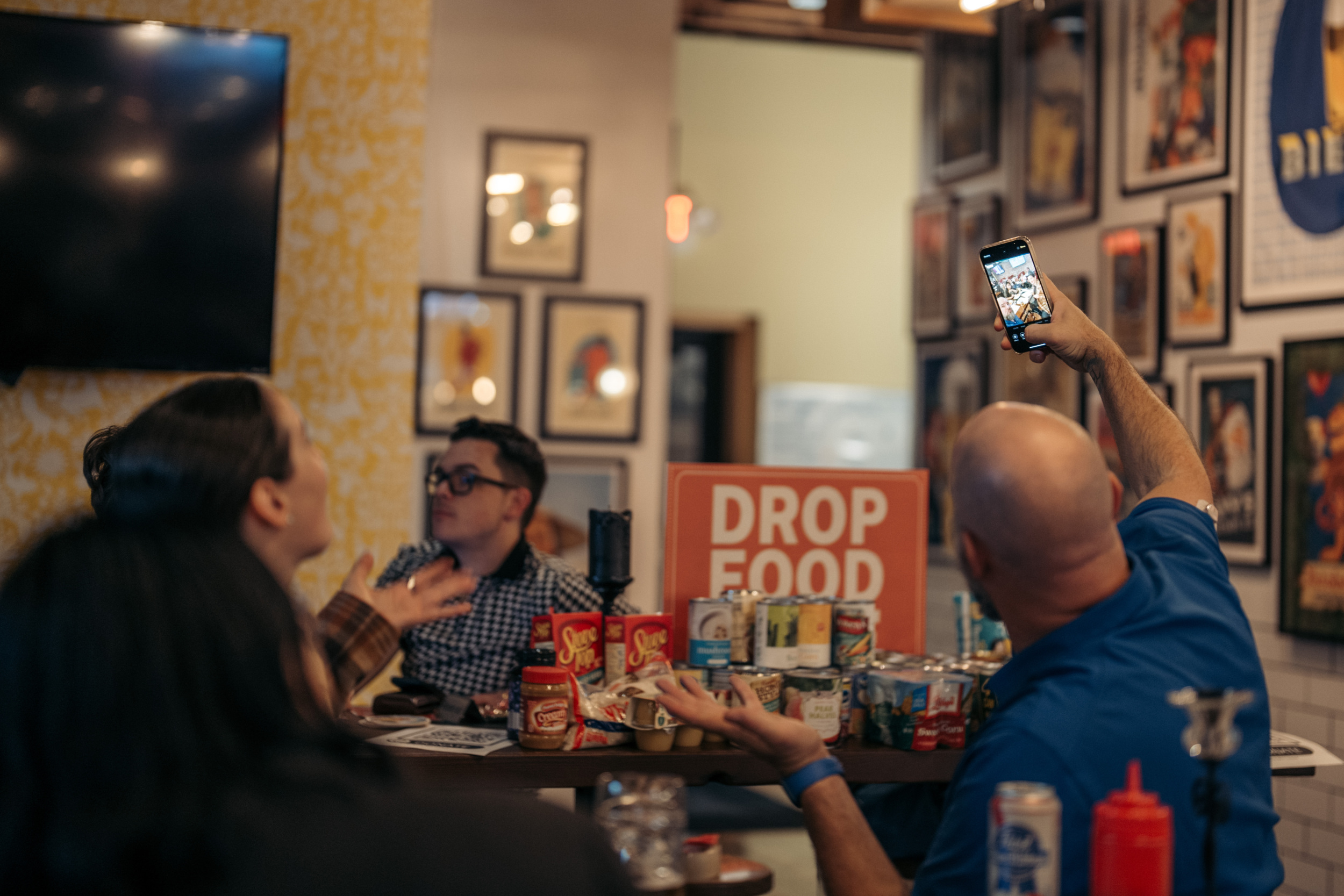 A man takes a selfie inside a restaurant. Food for donation are pictured in the background.